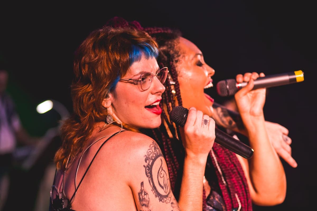 Duas vocalistas cantando lado a lado ao microfone sob luz quente de palco