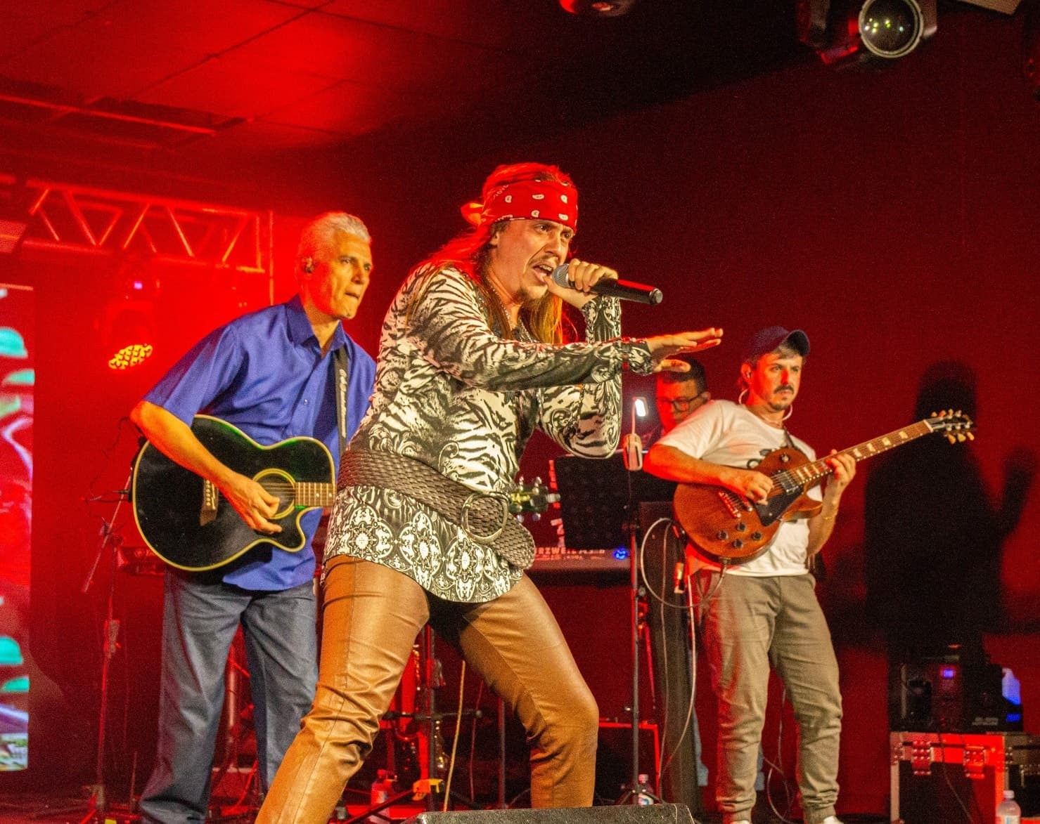 Vocalista de bandana vermelha à frente, com violão e guitarra ao fundo sob luz vermelha