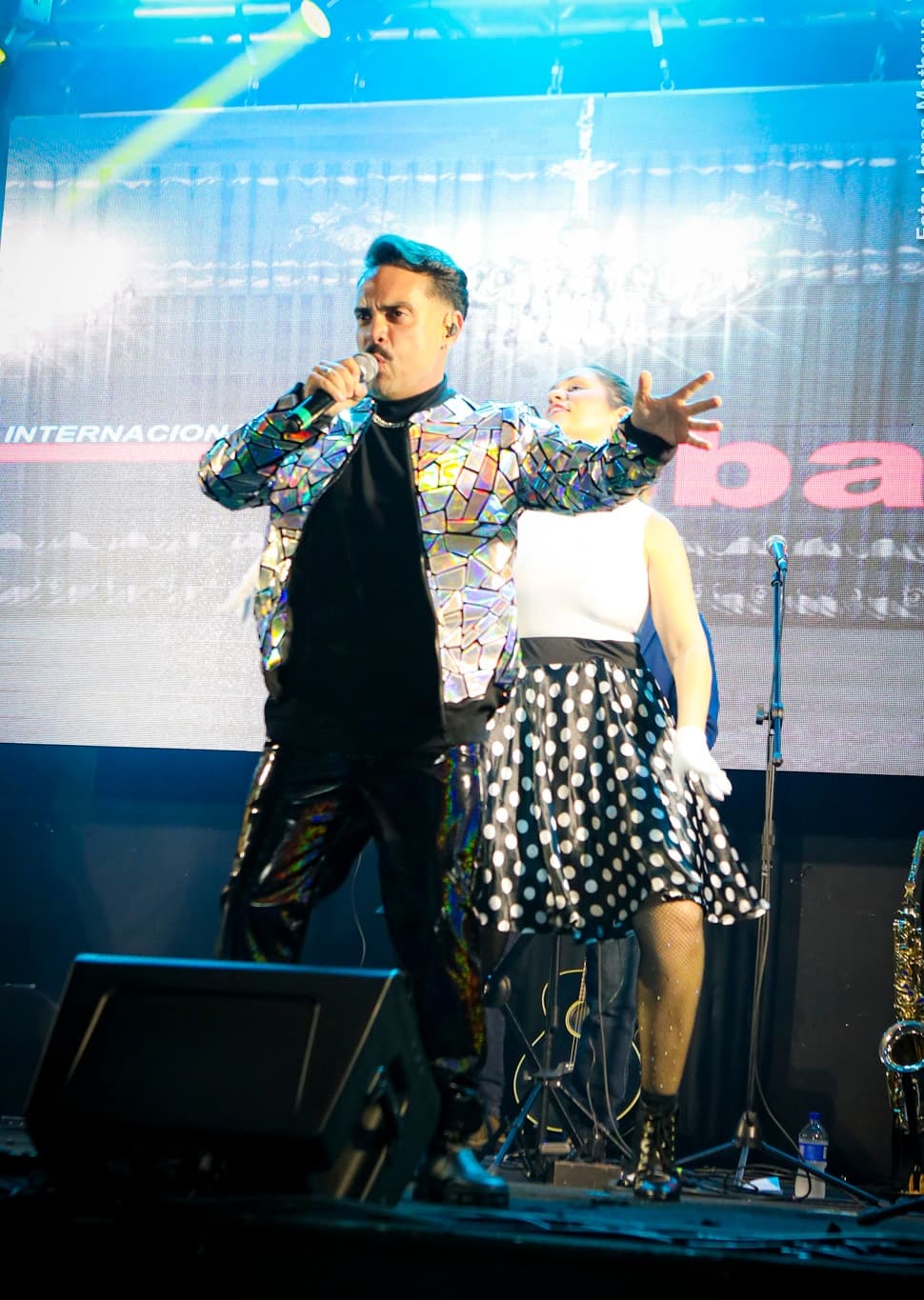 Vocalista de jaqueta espelhada e bailarina de saia de poá do bloco anos 50 diante do painel de LED