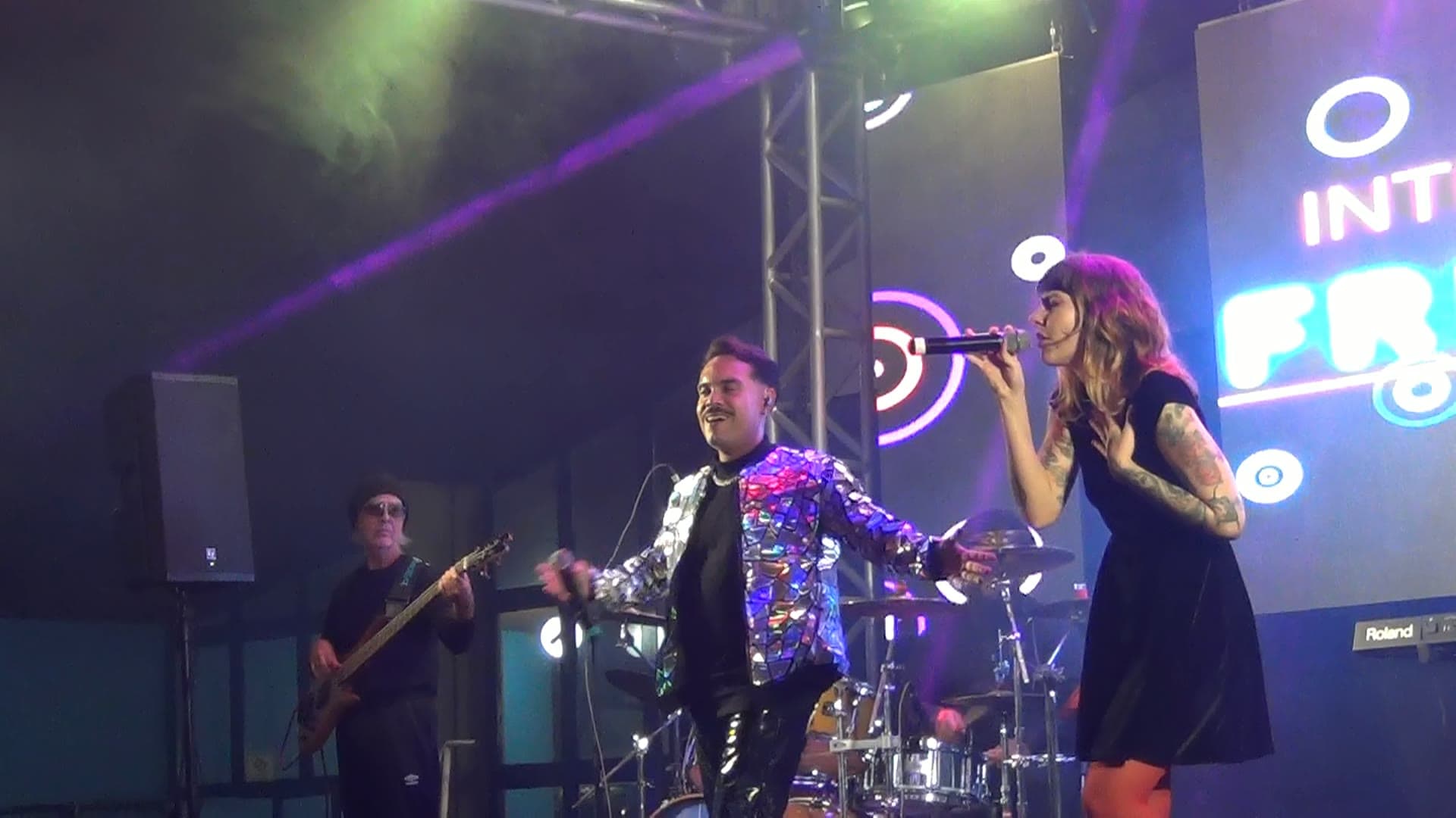 Vocalista de jaqueta espelhada e vocalista de vestido preto cantando lado a lado, com o baixista e a bateria atrás e o painel de LED aceso