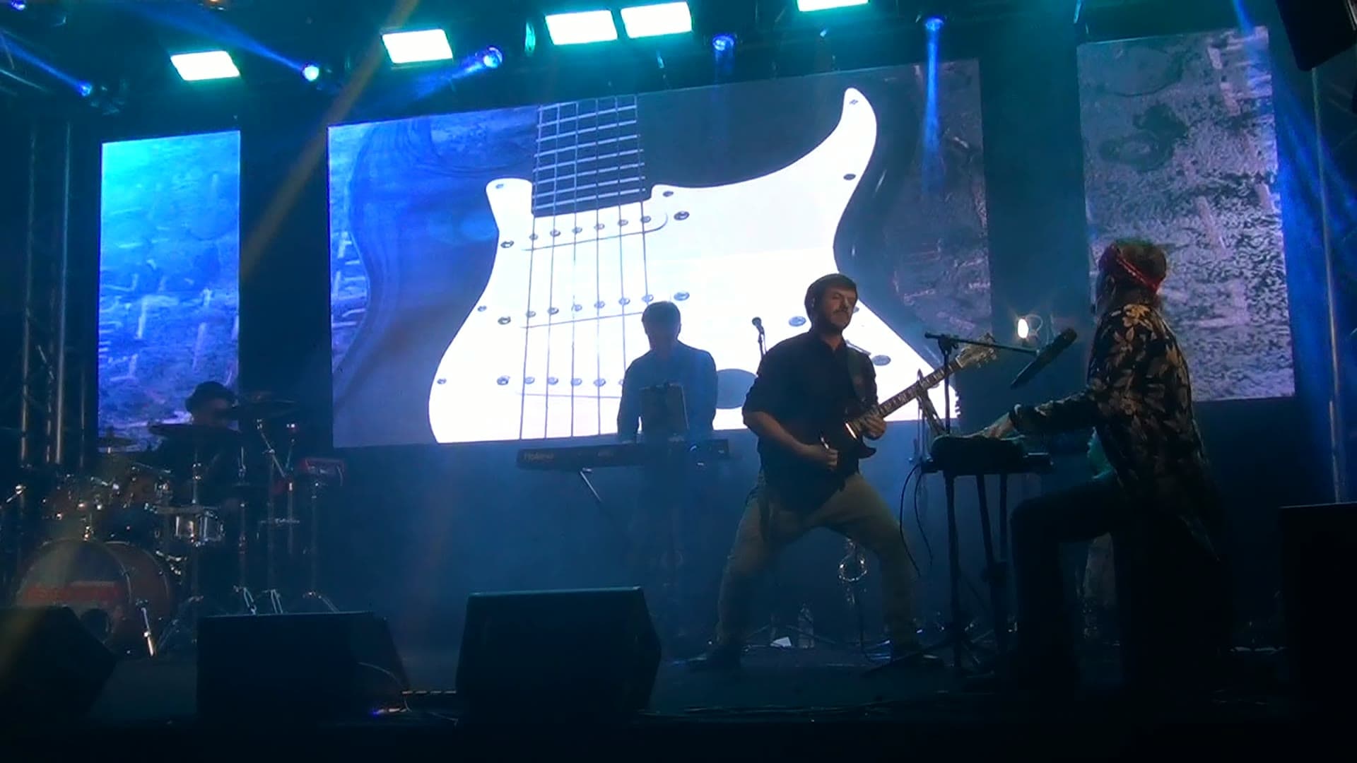 Guitarrista em solo sob luz azul, à frente de uma guitarra branca desenhada no painel de LED, com bateria e teclados ao redor