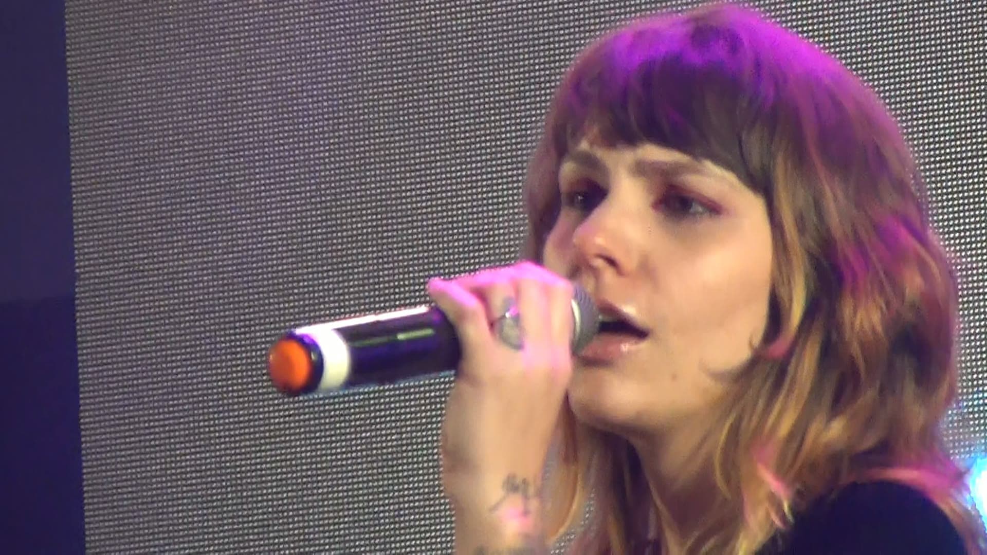 Vocalista cantando ao microfone em close, sob luz magenta, com o painel de LED ao fundo